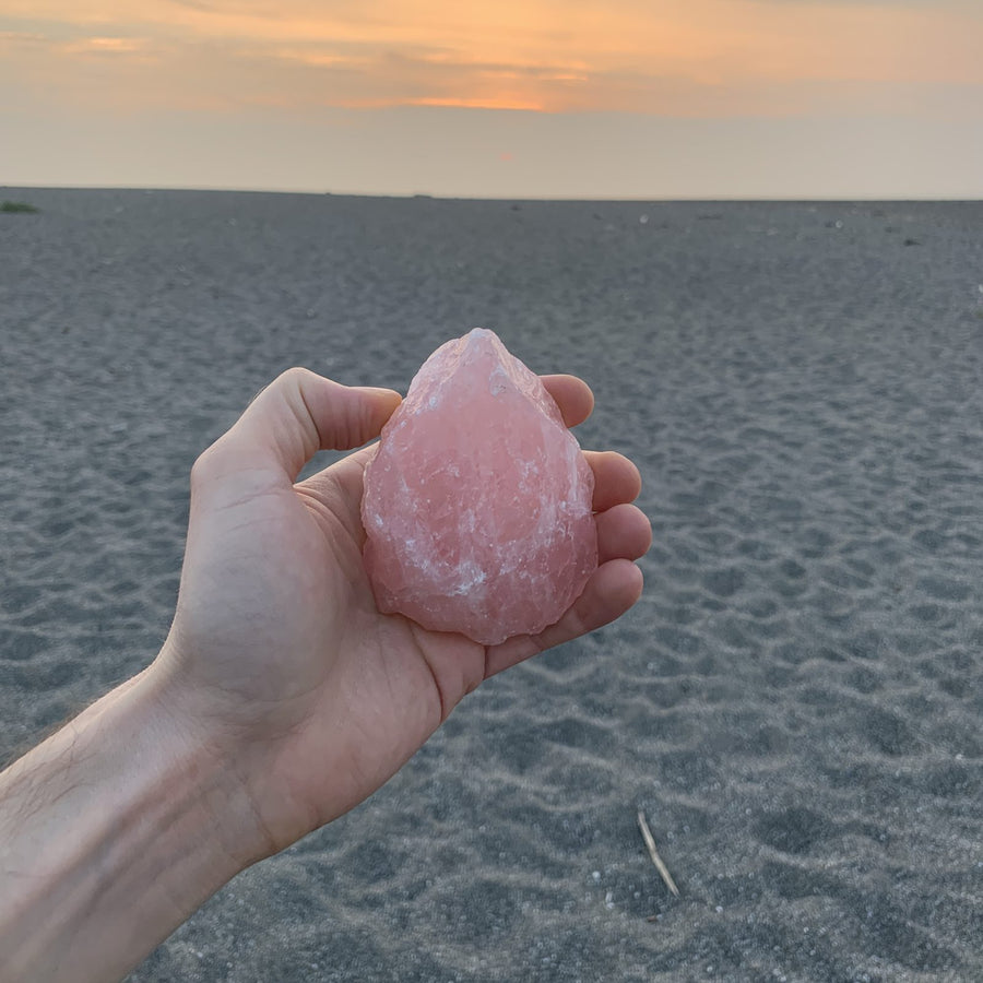 A hand holding a raw rose quartz point over gray sand at sunset