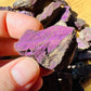 Held against neutral backdrop is a Purpurite raw crystal, associated with Focus, captured in natural daylight.
